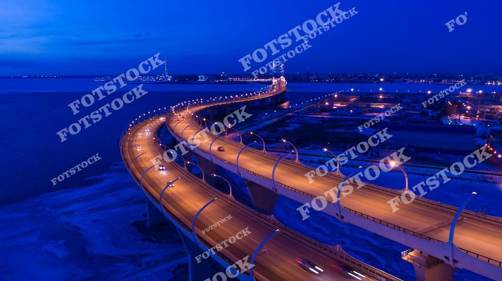 Фотография скоростного шоссе в Санкт-Петербург, Россия, сделанная в ночное время. На изображении показана современная автомагистраль мегаполиса, проходящая по мосту над водной поверхностью. Фотоснимок передаёт динамику ночного движения: потоки автомобилей, свет фар и художественную подсветку эстакад и дорожных развязок. Это фото подчёркивает развитую дорожную сеть Санкт-Петербурга и особенности современной транспортной архитектуры города. Иллюстрация отражает образ крупного мегаполиса с интенсивным трафиком и многоуровневыми скоростными дорогами.